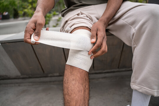 cropped view of african american man sitting on border bench and bandaging injured knee.