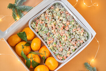 Traditional New Year and Christmas salad on holiday, in take way box. Menu food for delivery in the Coronavirus Pandemic. Tangerines and branches of the Christmas tree.