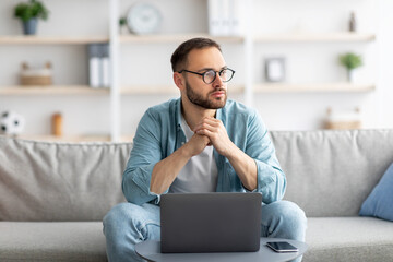 Serious young Caucasian guy sitting on couch in front of laptop computer at home