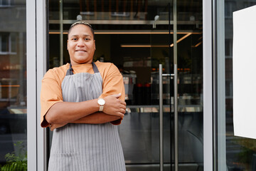 Horizontal medium portrait of young Black woman wearing apron standing relaxed with arms crossed at...
