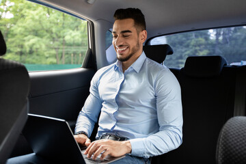 Cheerful middle eastern businessman working at car, using laptop