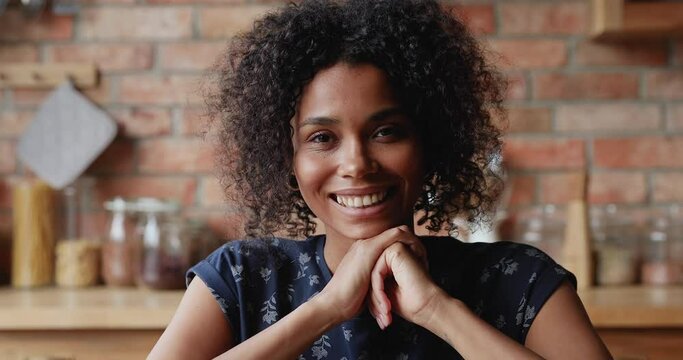 Young African woman having wide charming toothy smile look at camera giggling pose alone sit in kitchen at home. Positive female with natural curly black hair close up portrait. Natural beauty concept