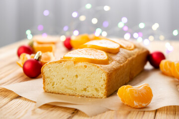 Loaf of tangerine cake with pieces of mandarin, red Christmas balls and lights on rustic wooden background. Tasty dessert for winter holidays. Merry Christmas and Happy New Year atmosphere.