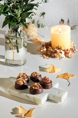 Chocolate pralines with walnut nuts on marble cutting board. Closeup on candy. Autumn arrangement with candle and ginkgo leaves. Natural sunlight, long shadows. Green eucalyptus twigs in glass bottle.