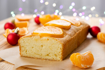 Moist tangerine pound cake on parchment on rustic wooden background with pieces of mandarin and Christmas lights bokeh. Delicious pastry for traditional English tea time on winter holidays.