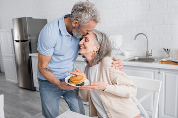 Mature man kissing smiling wife with tasty pancakes in kitchen.