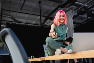 happy businesswoman with pink hair holding pen and notebook while sitting on desk and looking at...