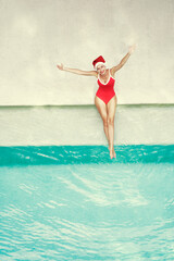 Enjoying christmas holidays and vacation. Top view of excited young woman in red swimsuit  and  santa claus hat rising hands up near swimming pool.