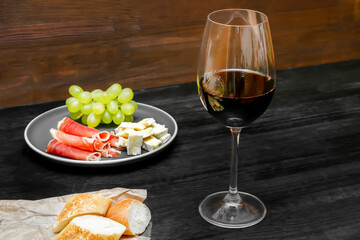 Red wine in glass, brie cheese, blue cheese, baguette, bread, grapes, jamon meat, salami on plate on black wooden table, alcohol drink with snacks, still life close up top view