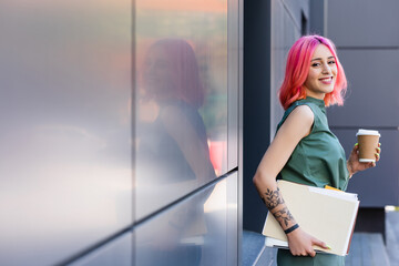 tattooed and happy businesswoman with pink hair holding folder, laptop and coffee to go outside.