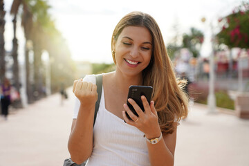 Laughing excited young woman watching her smartphone celebrating outdoor raising her fist up in...