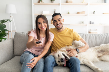 Happy young interracial couple sitting on couch with their funny dog and watching TV at home