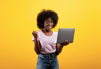 Euphoric african american lady celebrating success and holding laptop, raising clenched fist over...