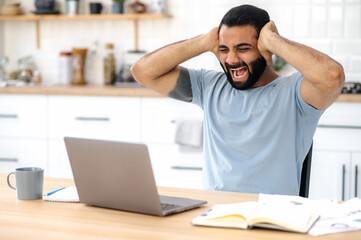 Indian man get a bad news. Angry annoyed Indian man, freelancer or designer, emotionally yells at a laptop, got bad news, failed project, lost, gesturing with hands, very irritated and frustrated