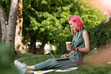 side view of happy woman with pink hair holding paper cup and using laptop on grass.