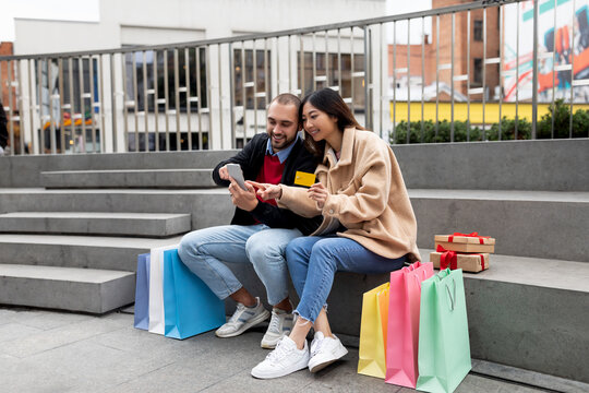 Young couple sitting on stairs with smartphone and credit card, shopping on web, surrounded with shopper bags and gifts