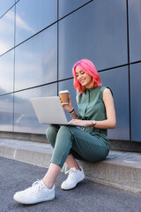 positive businesswoman with pink hair holding paper cup and using laptop outside.