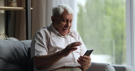 Laughing older adult male relax on couch look on cell screen overjoyed with good news in email message glad to get discount offer from web store. Aged grandpa feel excited winning online phone game