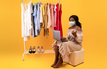 Remote shopping during covid. Young black lady in face mask sitting near clothing rack with laptop, showing thumb up