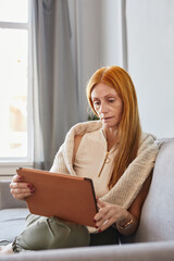Vertical portrait of adult red haired woman using digital tablet at home while sitting on sofa and studying online