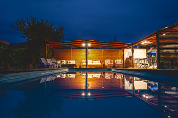 Obraz premium View of a big beautiful pool and wooden pavilion with barbecue in the countryside with beautiful lights and the blue sky in the night