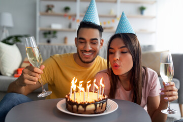 Young diverse couple in party hats celebrating birthday, blowing candles on festive cake, drinking...