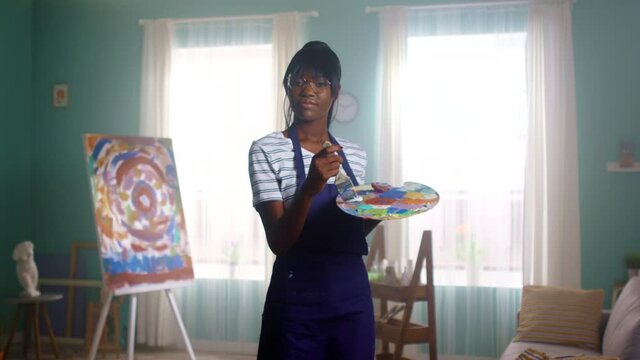 Young black woman, artist, is standing in painting studio, looking at camera confidently, playing with painting tools, mixing paint on palette, Slow motion.