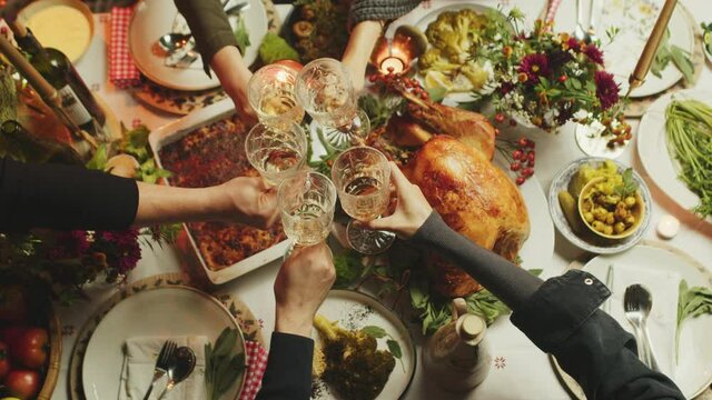People clink their glasses with white wine over plated and decorated thanksgiving table with big roasted turkey in the middle. Top view. Slow motion.