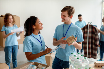 Young black woman checking charity list while group od volunteers making food packages for people in need