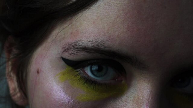 Close up on a woman's blue eyes, with yellow makeup in cyberpunk style. Young looking eyes with black eyeliner and yellow watercolor paint.