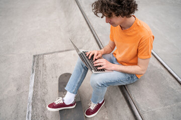young curly skateboarder typing on laptop in skate park.