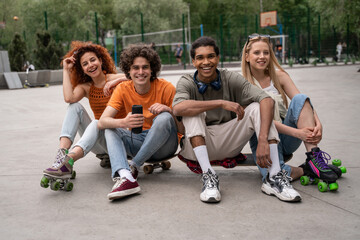 happy multiethnic skaters sitting on asphalt in skate park with portable music speaker.