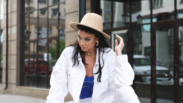 Young beautiful brunette in a white jacket and brown hat sits on the background of the building. Professional makeup and styling.Professional model beautifully posing for the camera