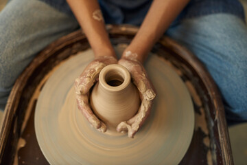 Hands of young female artisan touching clay jug while sitting by pottery wheel in her own workshop