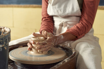 Schoolgirl in apron holding clay pot on rotating pottery wheel while attending handcraft class or...