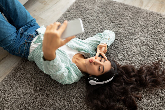 Top view of pretty Indian lady taking selfie on smartphone, lying on floor with headphones, listening to music at home