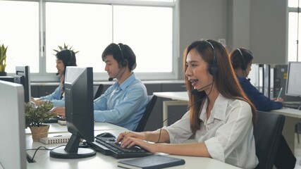 Fototapeta na wymiar Call center young employee working with headset, Smiling customer support operator team at work surrounded by colleagues working in the office, Helpdesk customer services support agent concept