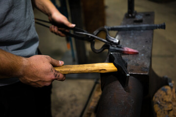 A blacksmith holds a hammer and a hot piece of metal with a pair of tongs on an anvil