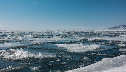 Ice drift on the river. Melted ice floes and loose snow on the surface of blue water. In the distance, against the background of the azure sky, tiny silhouettes of people walking are visible. Angara