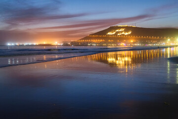 Fototapeta premium Beautiful evening view of the Atlantic ocean coast in Agadir, Morocco. With Kasbah mountain on the background with inscription in Arabian: Allah, The King, One's native land.