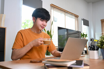 Happy man drinking coffee and using laptop computer at home in the morning.