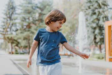Naklejka premium little boy is running in the street and playing with the water jets of a fountain spouting from the ground. child in blue T-shirt and white shorts is fooling around outdoors. lifestyle. space for text