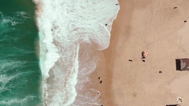 Static aerial drone showing endless wild sea waves breaking on the sandy coast line while people hang out on the beach. An umbrella is in the middle of the sand.