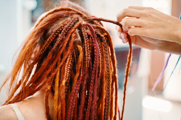 Woman hairdresser weaves girl ginger dreadlocks. Stylish therapy professional care concept. Close...