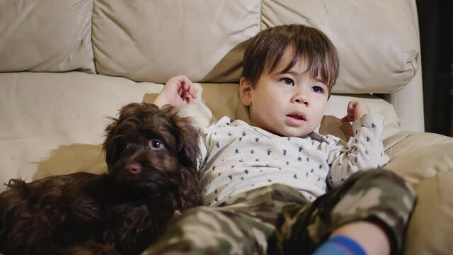 A kid of two years lies on the couch next to the puppy, watching TV