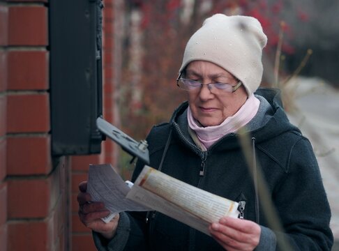 An elderly woman has opened a mailbox on the fence and is examining the correspondence she has received. A moment in the life of a pensioner.