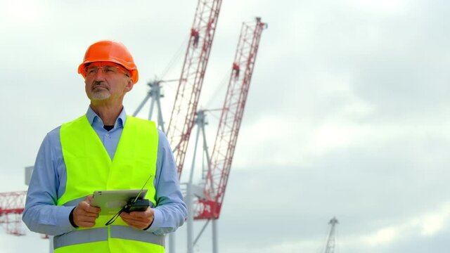 Sea cargo port industry. General manager with grey beard carries tablet and portable radio walking against high cranes in dockyard