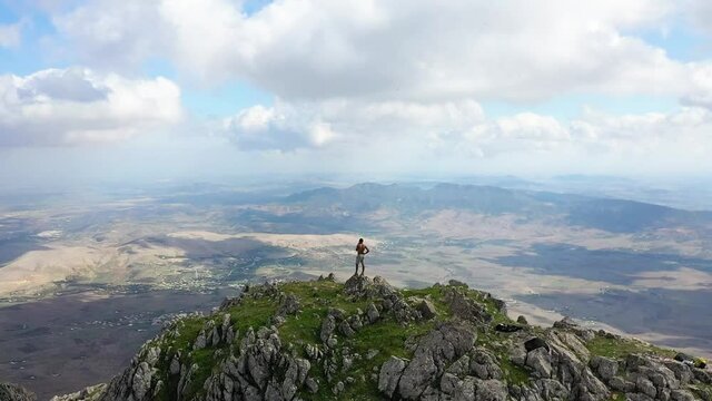 Circling drone shot of a local black man on top of the peak of Zaghouan in Tunisia, looking around to enjoy the amazing scenery and view with the epic and dramatic landscape below.
