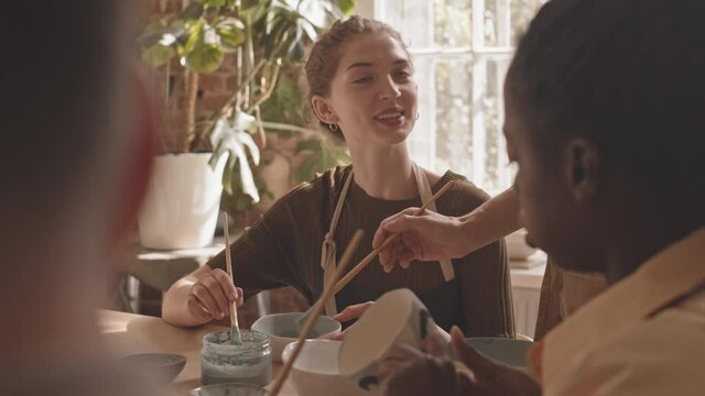 Slowmo chest up shot of young Caucasian woman attending group pottery masterclass, painting clay items together sitting at wooden table in cozy workshop