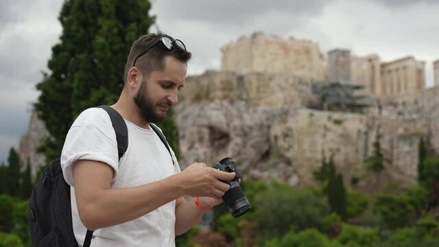 Young adult tourist stands among trees with camera, spends time on taking pictures of historical landmark. Traveler on sight of Acropolis in Athens. Tourism, traveling to ruins of ancient civilization